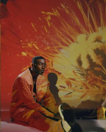 A stylish African-American man poses in a pink and orange knit jacket, seated against an abstract floral background with fashion-forward appeal.