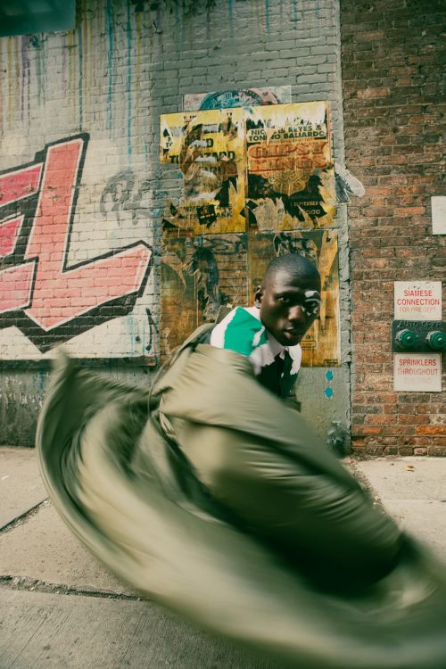 Urban street scene featuring a man in motion, partially wrapped in olive-green fabric, standing before a brick wall with graffiti.