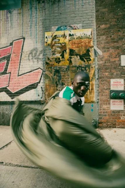 Urban street scene featuring a man in motion, partially wrapped in olive-green fabric, standing before a brick wall with graffiti.