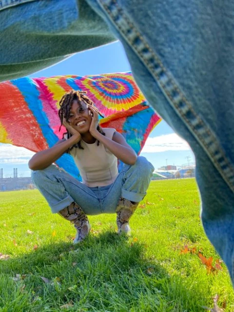 Smiling woman in jeans and snakeskin boots squats on a green grass lawn with a colorful tie-dye cloth, framed by legs in jeans.