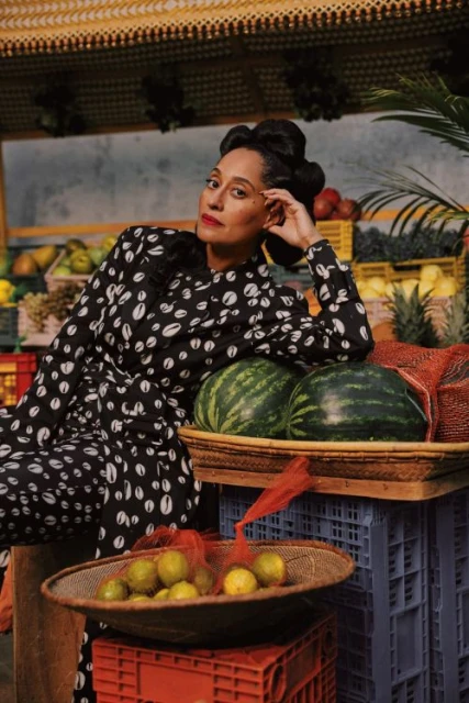 Tracee Ellis Ross poses in a patterned outfit with watermelons and lemons at a market stall, indoors, portrait orientation.