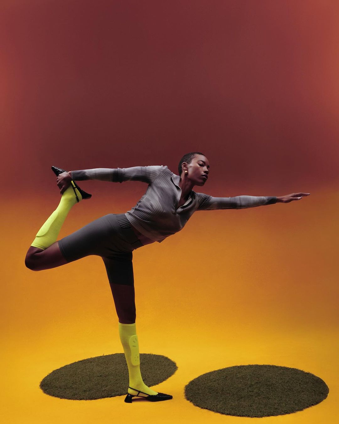 Athletic woman strikes yoga pose on ochre floor with green mats. She wears gray activewear and bright yellow socks with black flats.