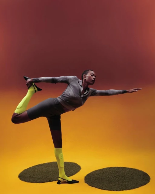 Athletic woman strikes yoga pose on ochre floor with green mats. She wears gray activewear and bright yellow socks with black flats.