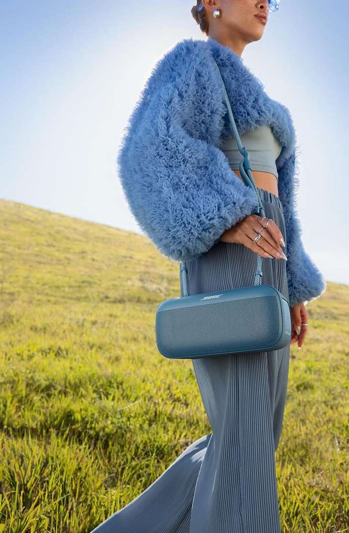 Woman in blue outfit carrying a speaker in an outdoor setting with grassy hills and a blue sky on a sunny day.