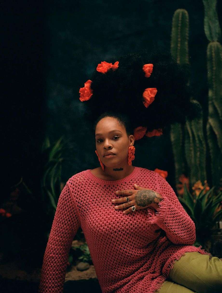 Woman with large Afro and orange flowers sits against cactus and plant background, wearing a pink sweater and green pants.
