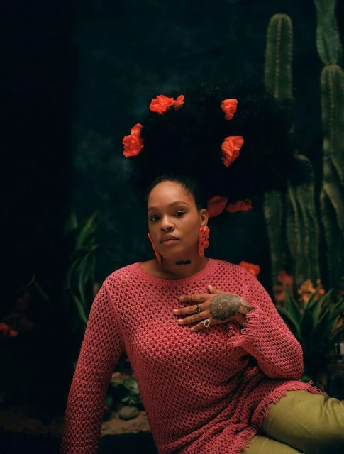 Woman with large Afro and orange flowers sits against cactus and plant background, wearing a pink sweater and green pants.