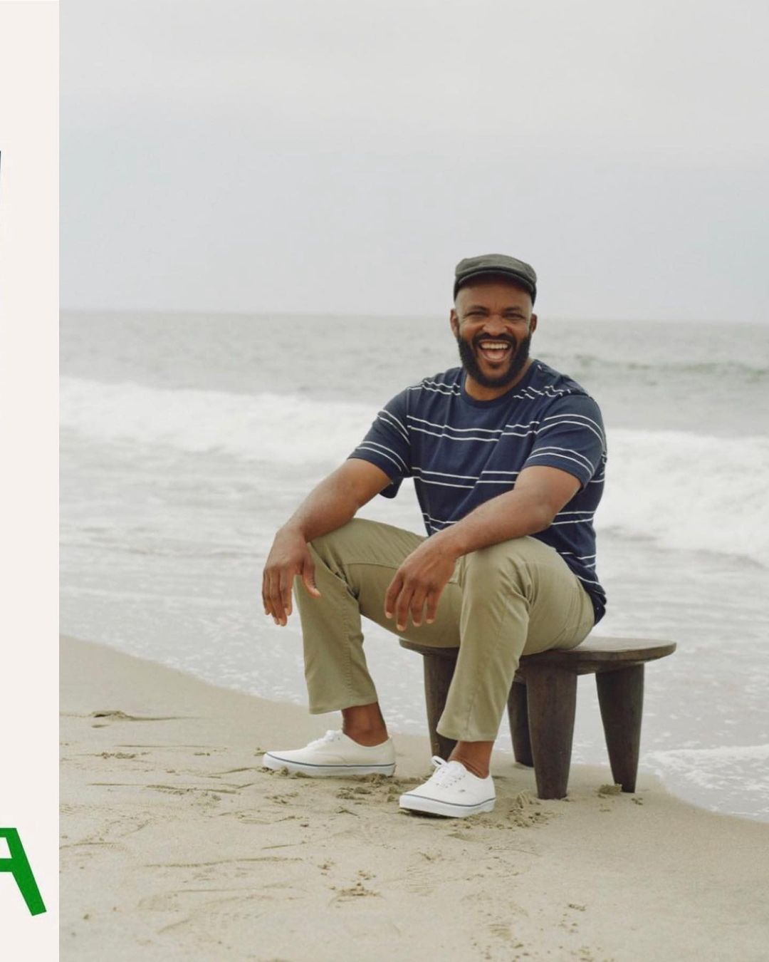 A smiling man with a beard sits on a stool on a beach with ocean waves, wearing a striped shirt, tan pants, a cap, and white sneakers.