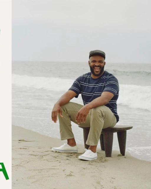 A smiling man with a beard sits on a stool on a beach with ocean waves, wearing a striped shirt, tan pants, a cap, and white sneakers.