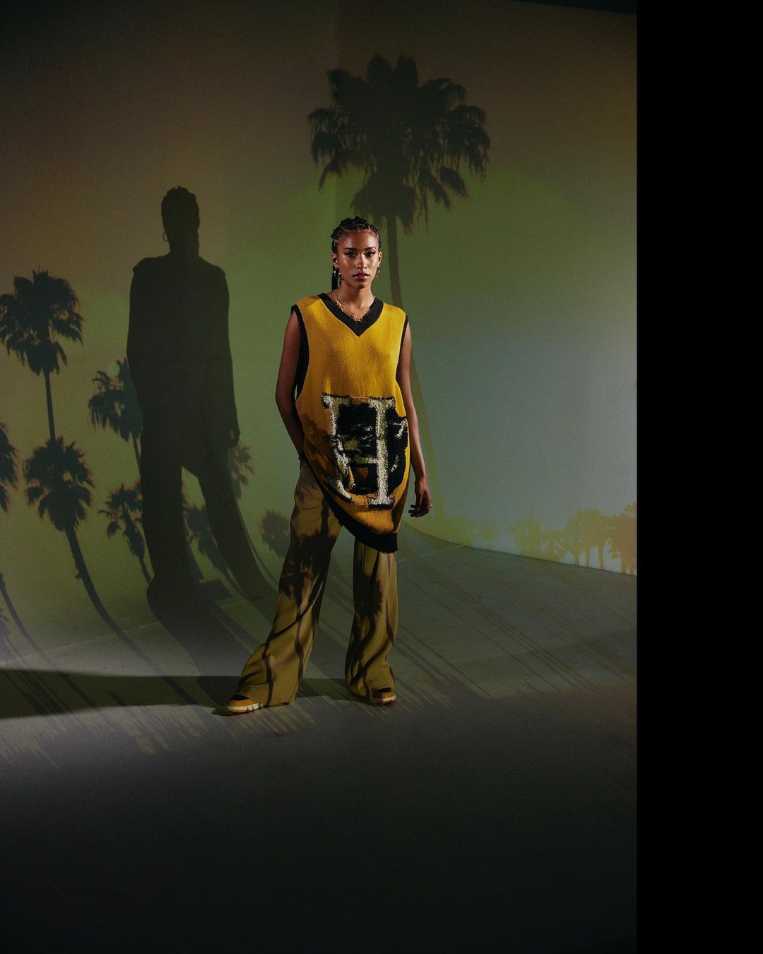 Black woman wearing a yellow sweater vest and patterned trousers standing against a projected palm tree backdrop.