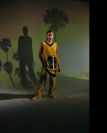 Black woman wearing a yellow sweater vest and patterned trousers standing against a projected palm tree backdrop.