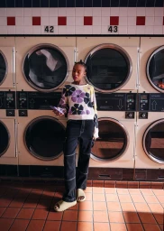 Black woman wearing a floral sweater and dark pants holding a phone inside a commercial laundromat