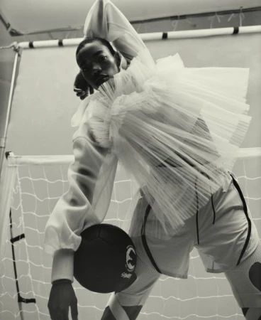 Black man wearing a ruffled sheer white top and holding a dark ball in front of a sports net background
