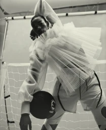 Black man wearing a ruffled sheer white top and holding a dark ball in front of a sports net background