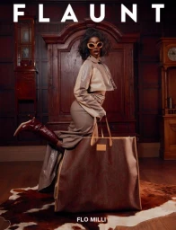Flo Milli wearing sunglasses and brown coordinates, posing playfully with a giant tote bag in a wood-paneled room by Mark Clennon.