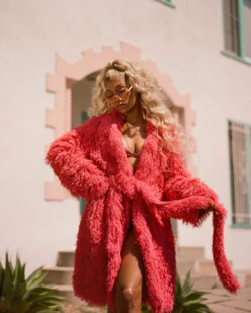 Woman with long blonde hair wearing a bright pink textured coat and tinted sunglasses standing outside a light-colored building.