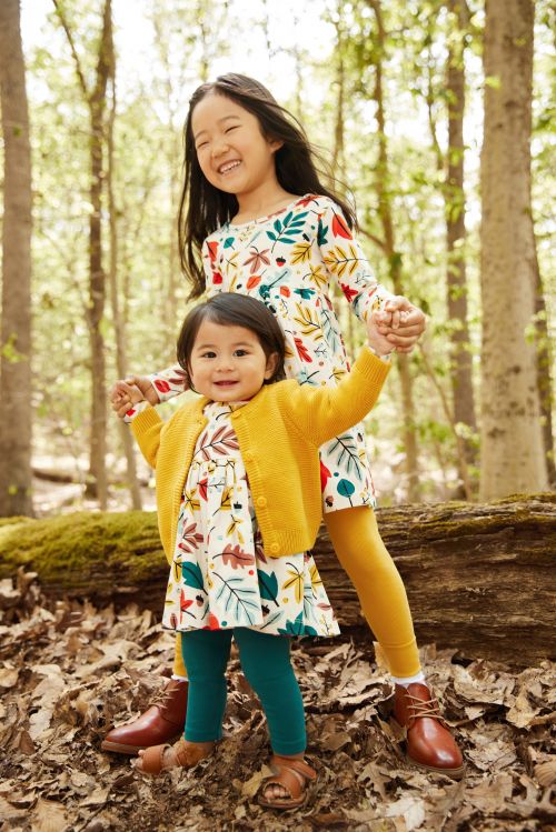 Two young children, an Asian girl and a toddler, hold hands in a forest setting. The older girl smiles in a leaf-patterned dress.