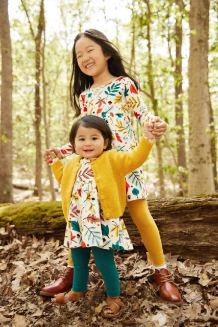 Two young children, an Asian girl and a toddler, hold hands in a forest setting. The older girl smiles in a leaf-patterned dress.