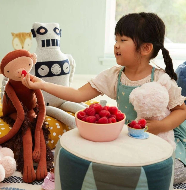 A young girl shares raspberries with a monkey plush toy, with other plush toys nearby, in a bright, inviting indoor setting.