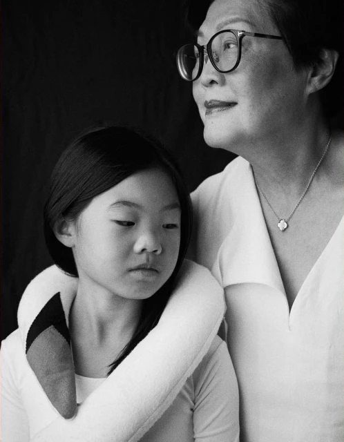 Black and white portrait of an Asian child and senior woman with short hair and glasses, wearing white, against a dark background.
