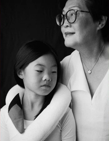 Black and white portrait of an Asian child and senior woman with short hair and glasses, wearing white, against a dark background.