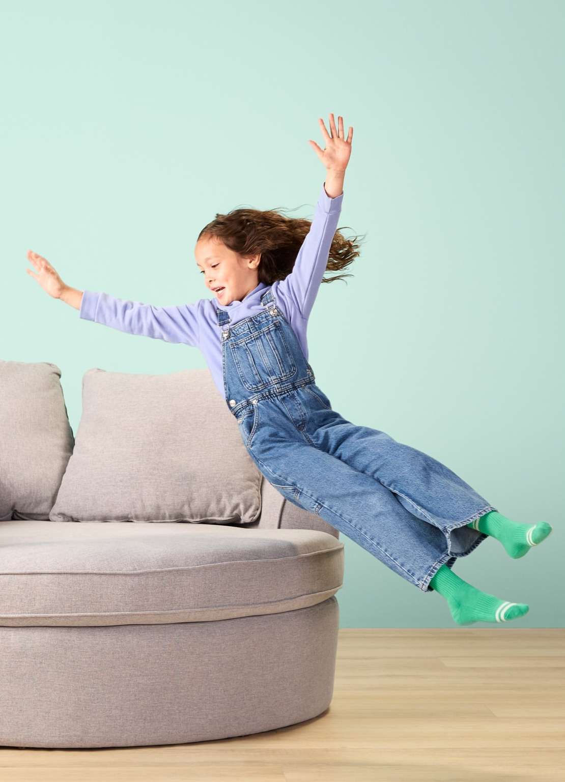 A young girl with curly hair is jumping off a gray rounded sofa, arms outstretched, wearing denim overalls and green socks in a light teal room.