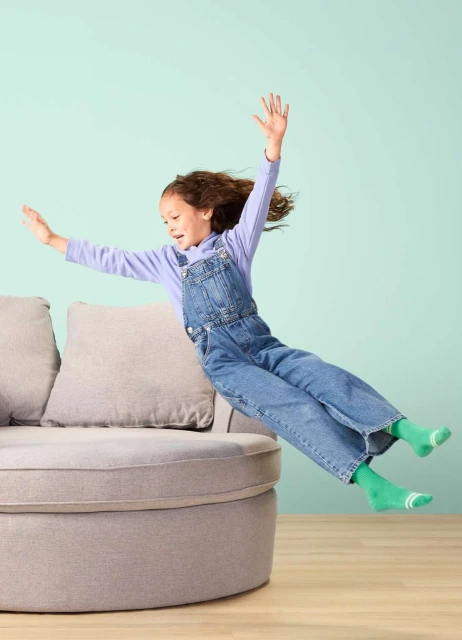 A young girl with curly hair is jumping off a gray rounded sofa, arms outstretched, wearing denim overalls and green socks in a light teal room.