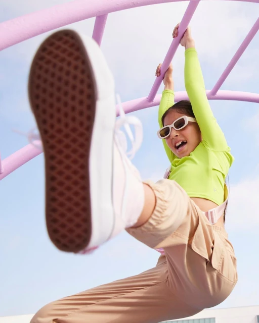 A child hangs upside down from a purple climbing frame, wearing a lime green shirt, beige pants, and white sneakers against a blue sky.