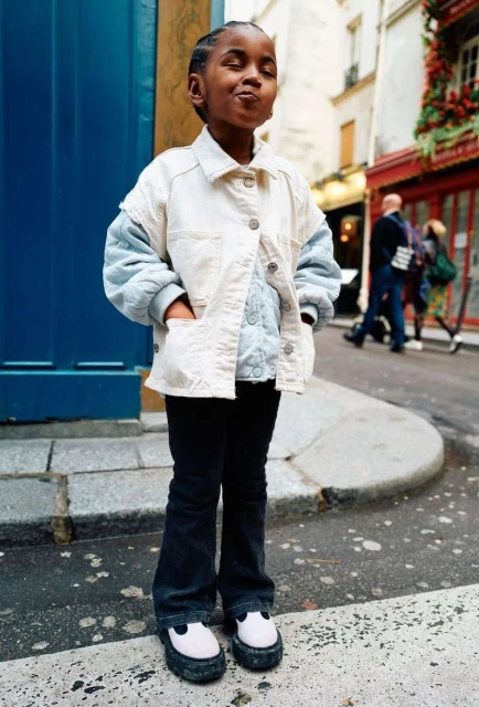 Young child poses in a cream jacket, blue sweater, and black pants in an urban setting. The image features a classic, youthful fashion style.