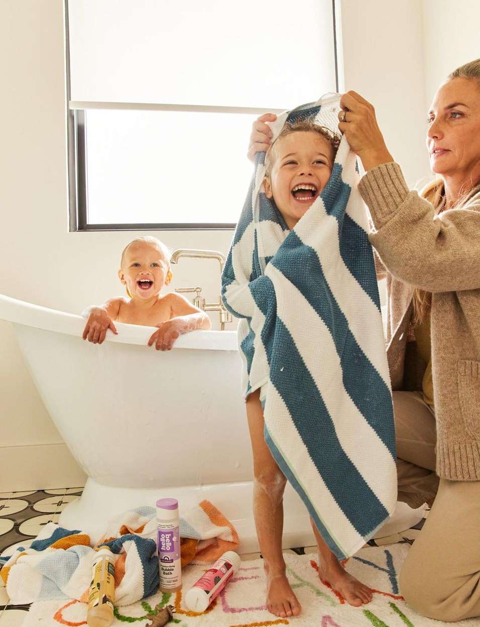 Two children and an adult in a bright bathroom after bath time, featuring a white bathtub, patterned rug, and striped towel.