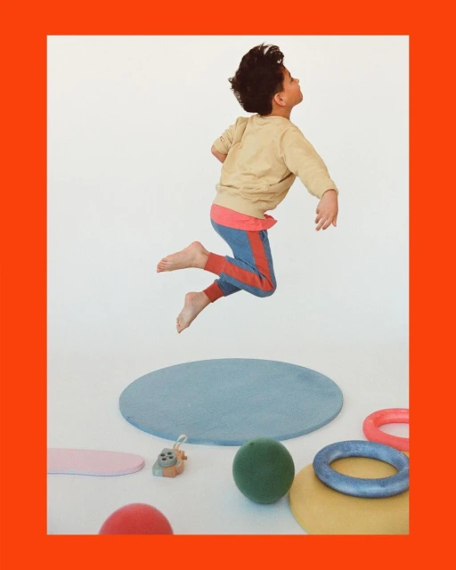 A young boy wearing a beige sweatshirt and blue pants with a red stripe leaps in the air inside a photo studio with toys and props.