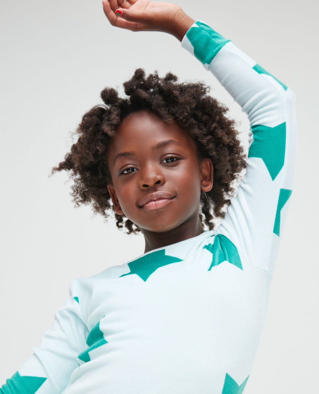 Portrait of a smiling Black child with curly hair, wearing a star-patterned shirt against a neutral background, looking at the camera.