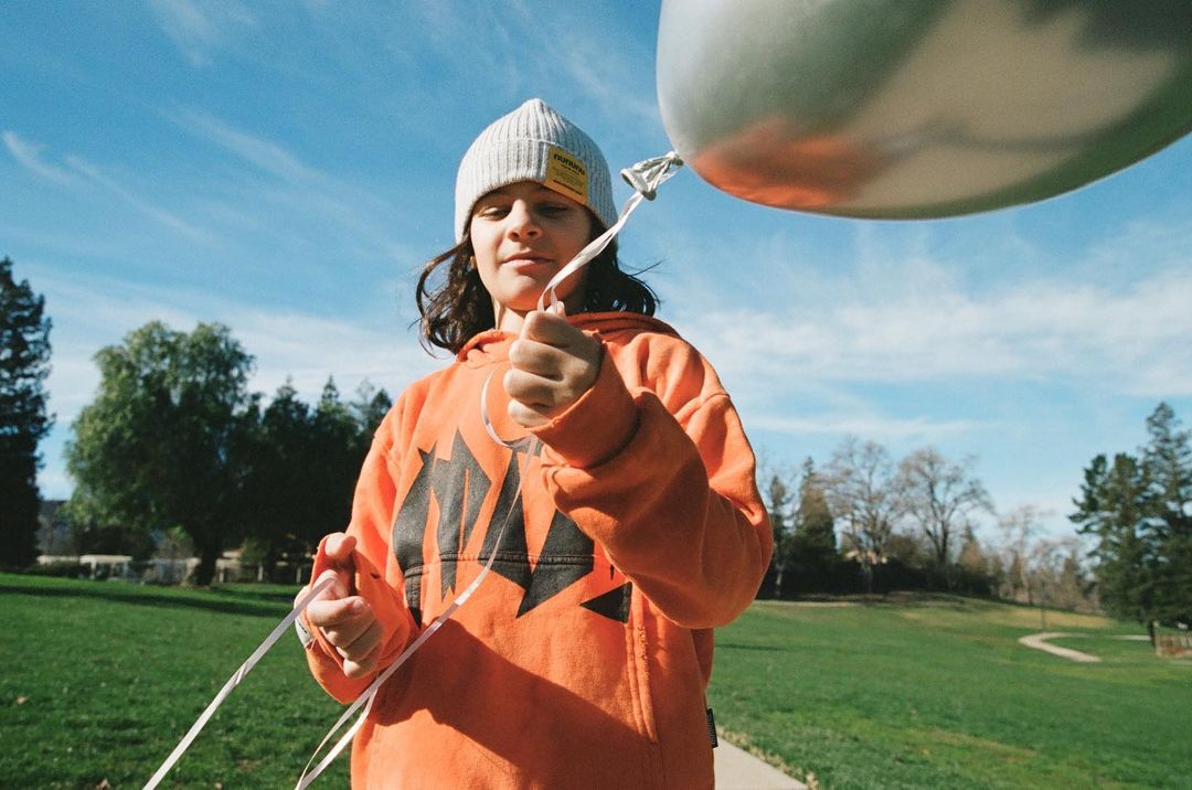Person in orange hoodie holds a large silver balloon outdoors in a grassy park-like setting under a partly cloudy blue sky.