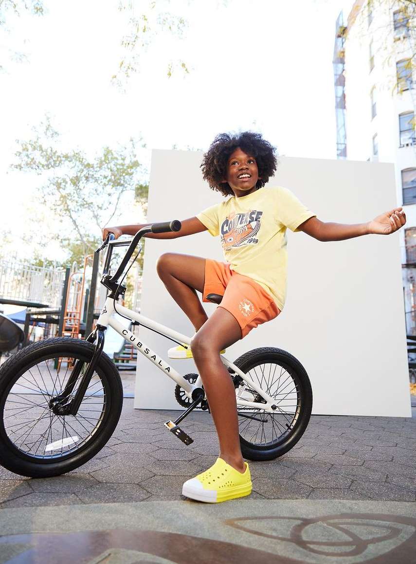 Young boy with afro hair smiling while riding a BMX-style bicycle outdoors near a concrete building on a sunny day.