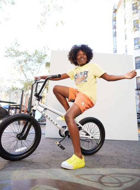 Young boy with afro hair smiling while riding a BMX-style bicycle outdoors near a concrete building on a sunny day.