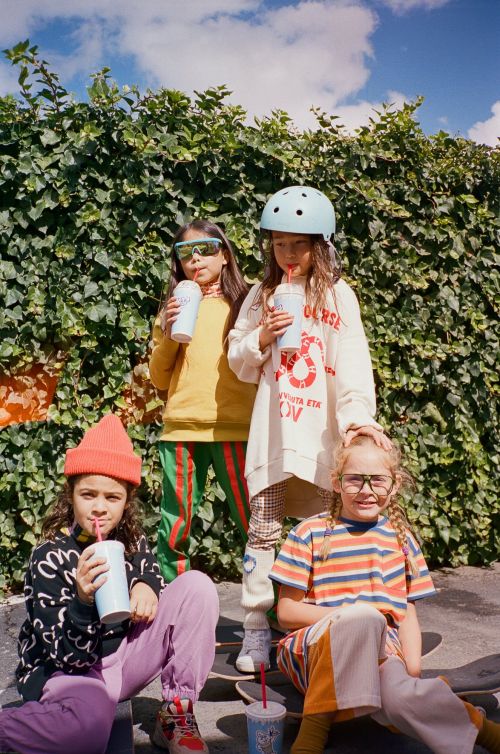 Four young girls in colorful casual clothes are sitting on a skateboard ramp outdoors, drinking from cups with straws, with green foliage in background.