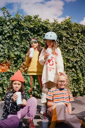 Four young girls in colorful casual clothes are sitting on a skateboard ramp outdoors, drinking from cups with straws, with green foliage in background.