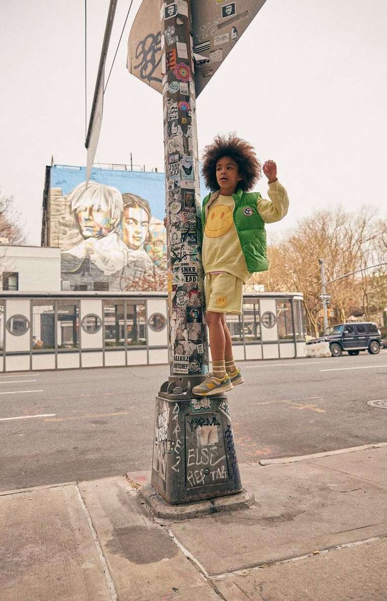 Young child in yellow smiley face sweatshirt and green vest climbs a sticker-covered pole in an urban street setting.