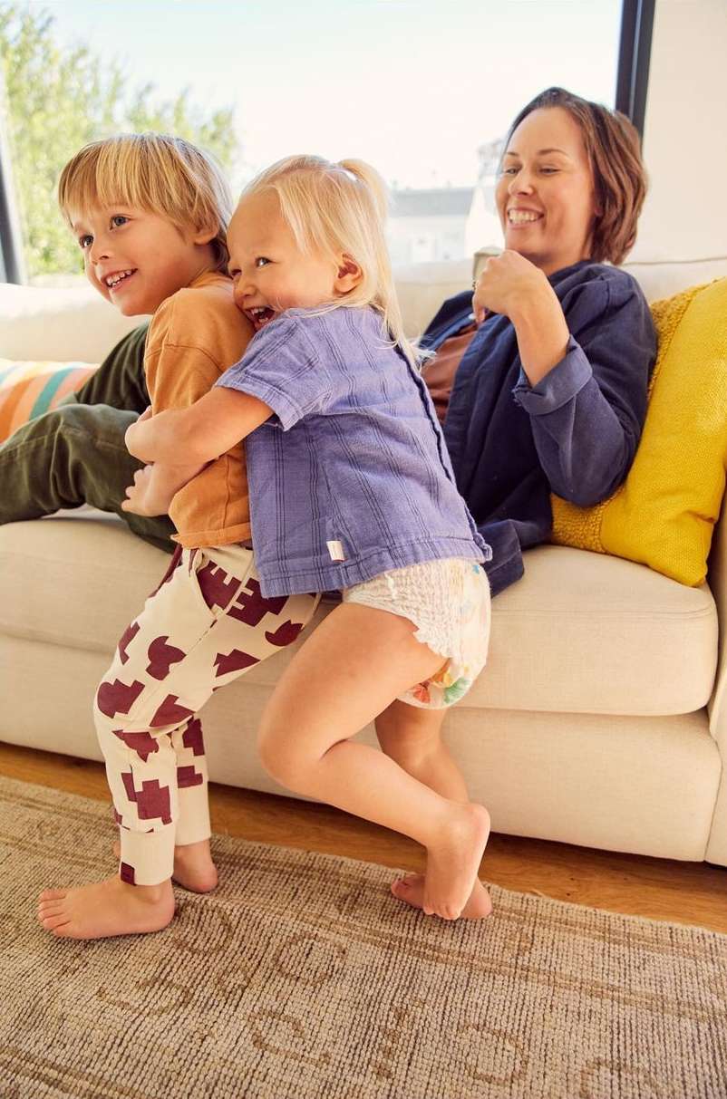 Two young children hug tightly while a mother smiles on a sofa indoors. The children, one in cow print pants, show joy and affection.