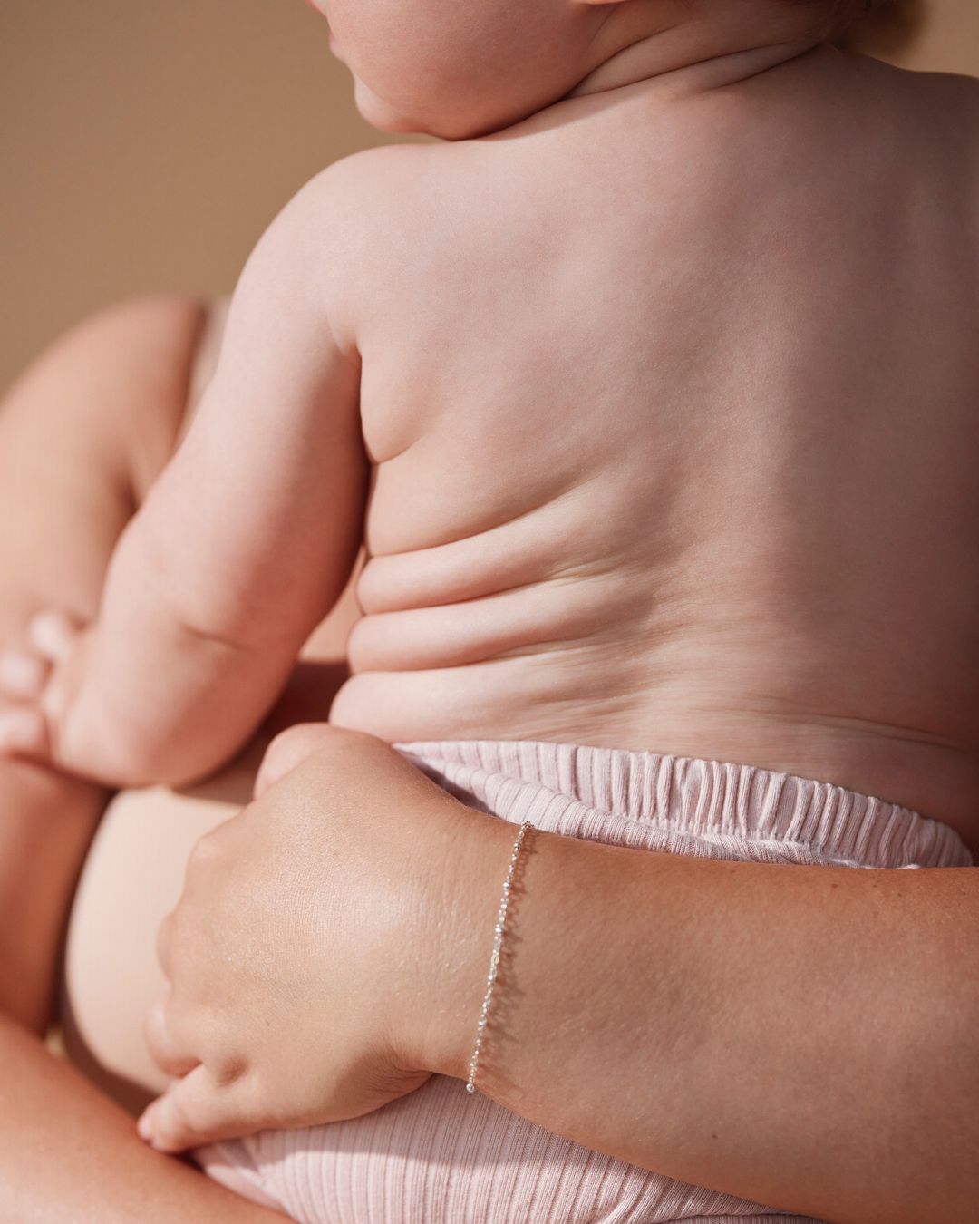 Toddler with light skin and some skin rolls sits in their mother's lap and looks away, while the mother's arm is visible.