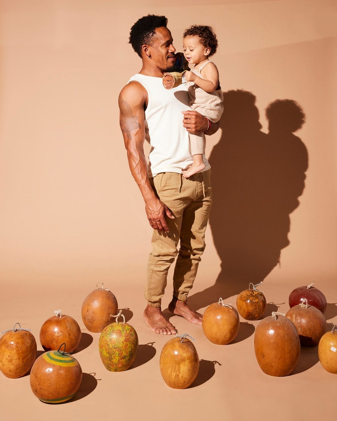 A muscular man holds a toddler while standing barefoot on a tan surface surrounded by gourds in a brightly lit indoor setting.