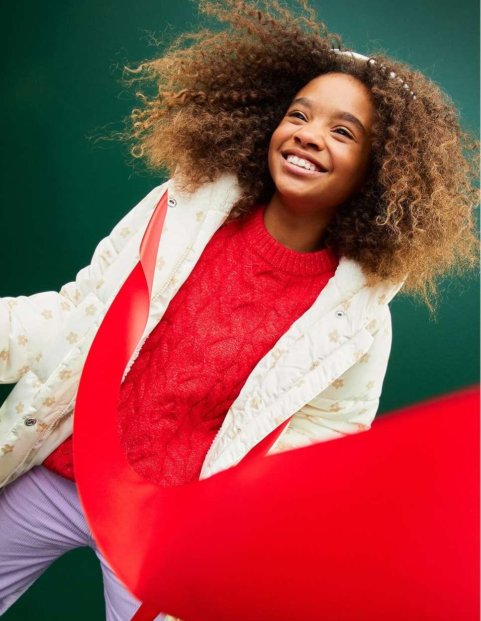 A joyful young girl with curly hair smiles against a green backdrop, wearing a cream quilted jacket, red sweater, and lavender pants.