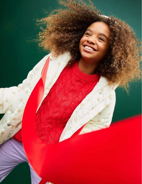 A joyful young girl with curly hair smiles against a green backdrop, wearing a cream quilted jacket, red sweater, and lavender pants.
