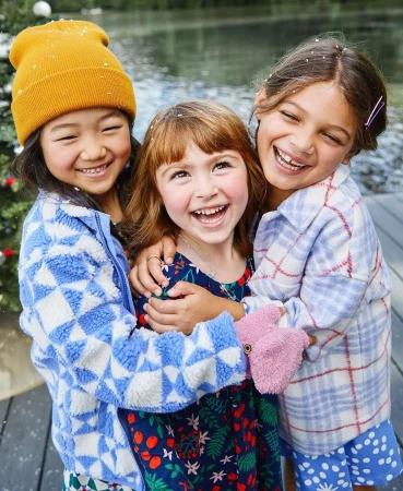 Three smiling young girls embrace outdoors in a close-up portrait; one wears a beanie, one has a floral dress, and the third a plaid shirt.