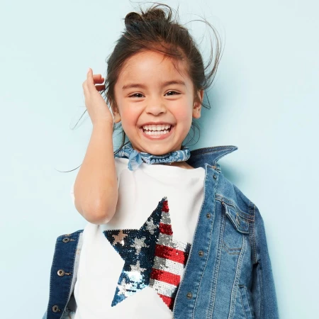 Smiling child with hair in a top knot wearing a white star t-shirt and denim jacket against a light blue background.