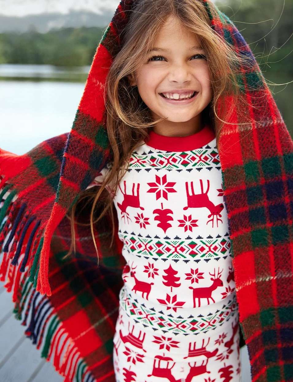 A smiling girl wrapped in a red and green plaid blanket wears reindeer pajamas outdoors by a lake, creating a cozy, festive scene.