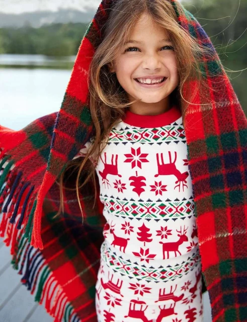 A smiling girl wrapped in a red and green plaid blanket wears reindeer pajamas outdoors by a lake, creating a cozy, festive scene.