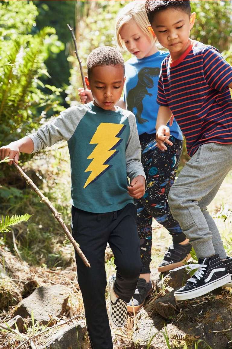 Three children hiking on a wooded path, wearing casual clothes and sneakers. One child carries a stick as they walk on rocks and tree roots.