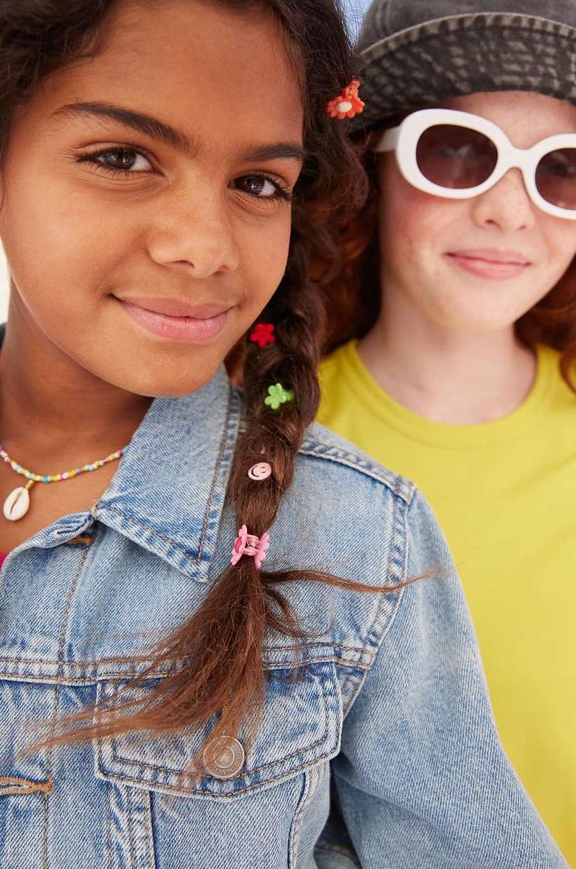 Two young girls smiling, one with a braided hairstyle adorned with colourful clips, wearing a denim jacket, and another with sunglasses.