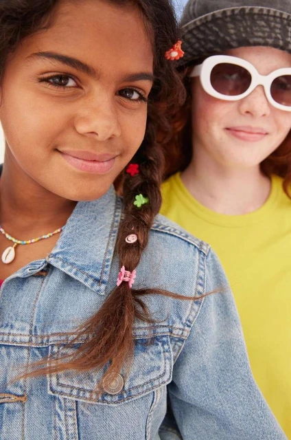 Two young girls smiling, one with a braided hairstyle adorned with colourful clips, wearing a denim jacket, and another with sunglasses.