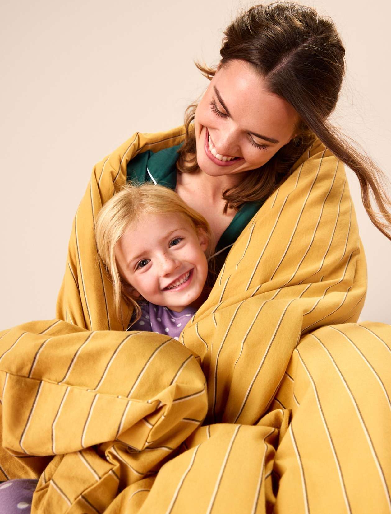 A cheerful mom and daughter are smiling wrapped in a yellow striped blanket, creating a cozy indoor portrait on a neutral background.
